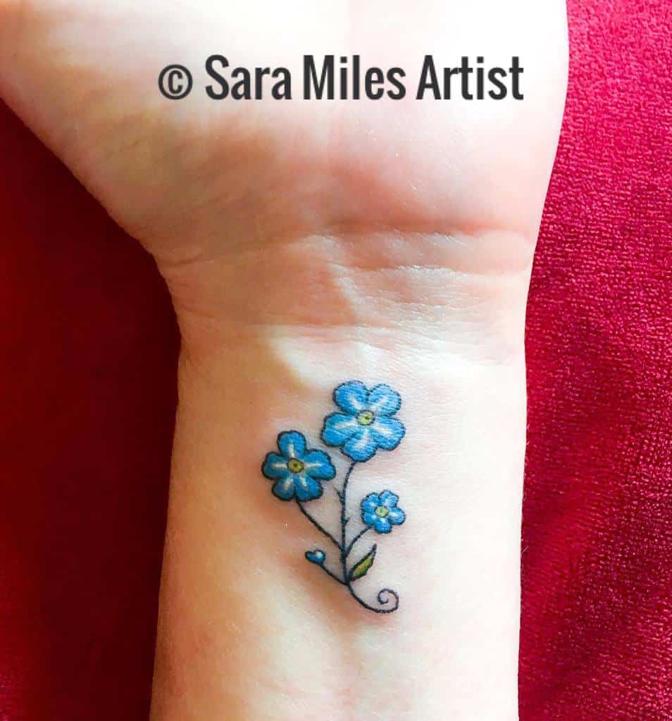A charming tattoo of three blue forget-me-nots with delicate swirling stems, beautifully inked on the wrist