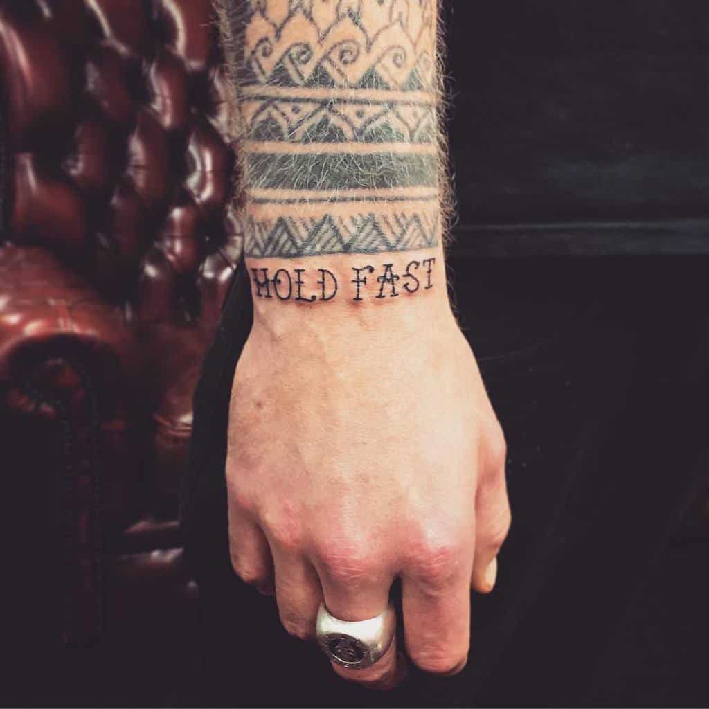 Close-up of a hand with a "HOLD FAST" tattoo and geometric patterned arm tattoo