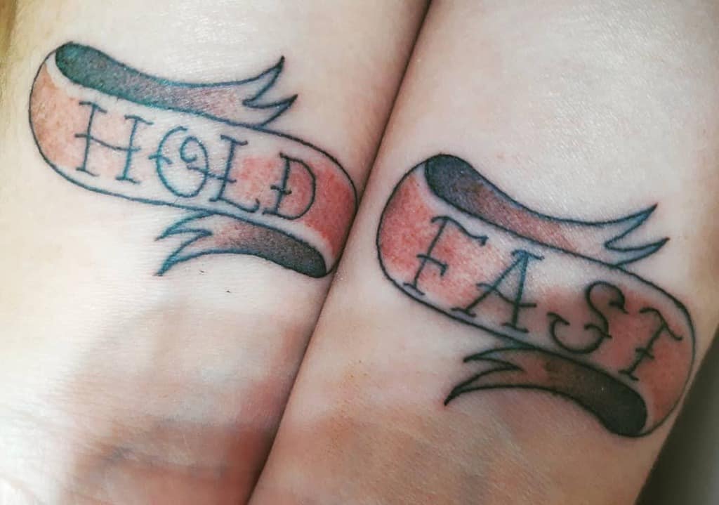 Minimalistic wrist tattoos featuring the phrase 'Hold Fast' on ribbon banners with a subtle shaded design