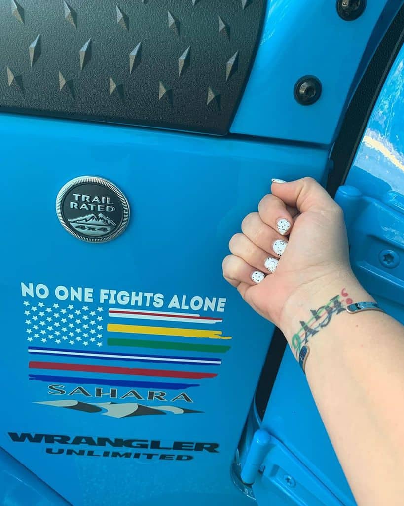 Wrist tattoo of ';IGY6' with a colorful design, shown against a blue Jeep Wrangler door with 'No One Fights Alone' text