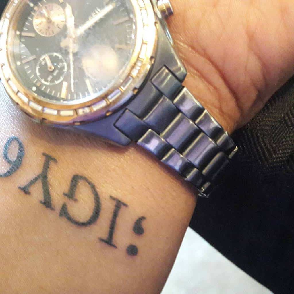 Minimalist wrist tattoo of ';IGY6' in black ink, shown with a silver and gold wristwatch