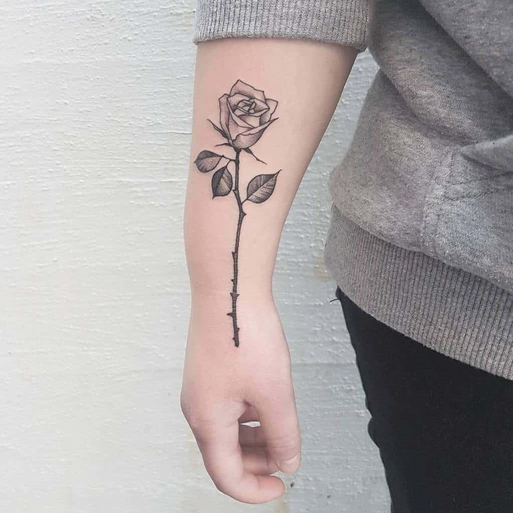 Elegant forearm tattoo of a single rose with a long stem, detailed leaves, and subtle thorns, symbolizing beauty and strength