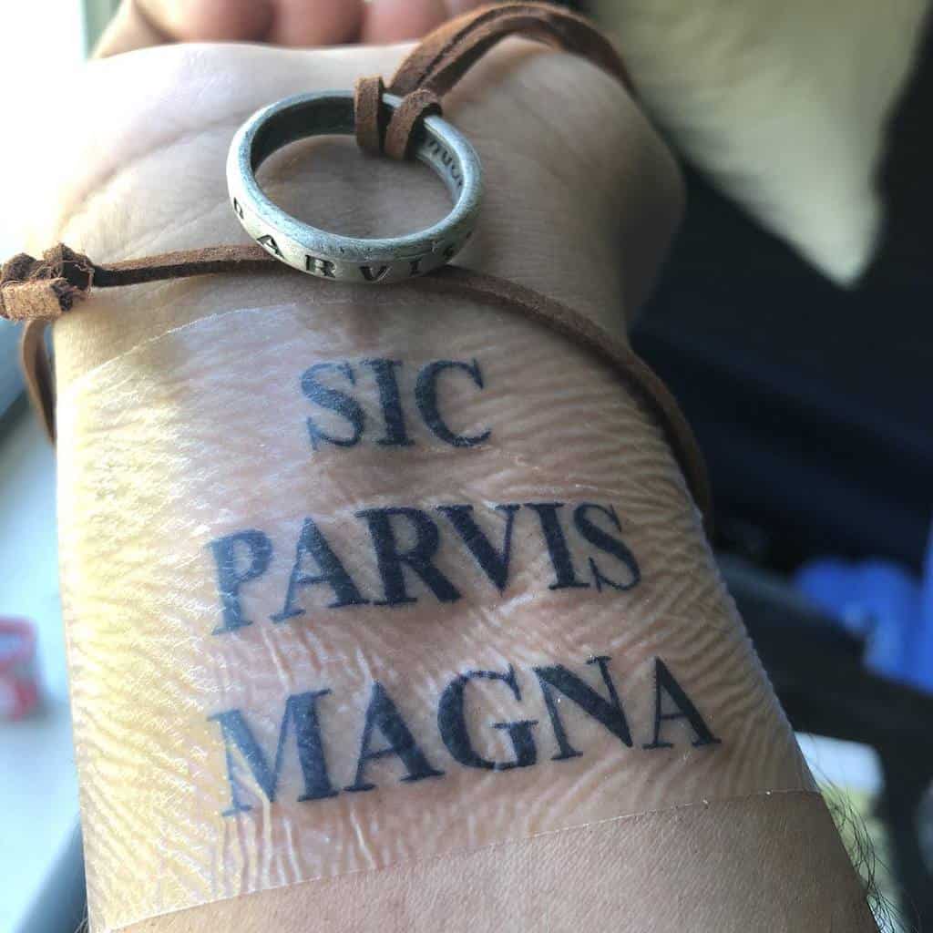 Tattooed wrist with the phrase "Sic Parvis Magna" and a ring on a leather strap