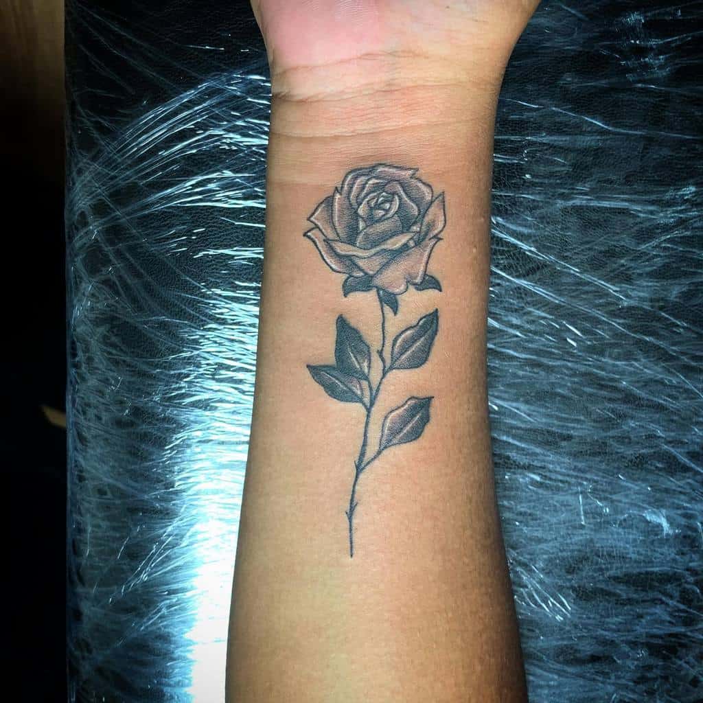 Forearm tattoo of a detailed, shaded rose with stem and leaves