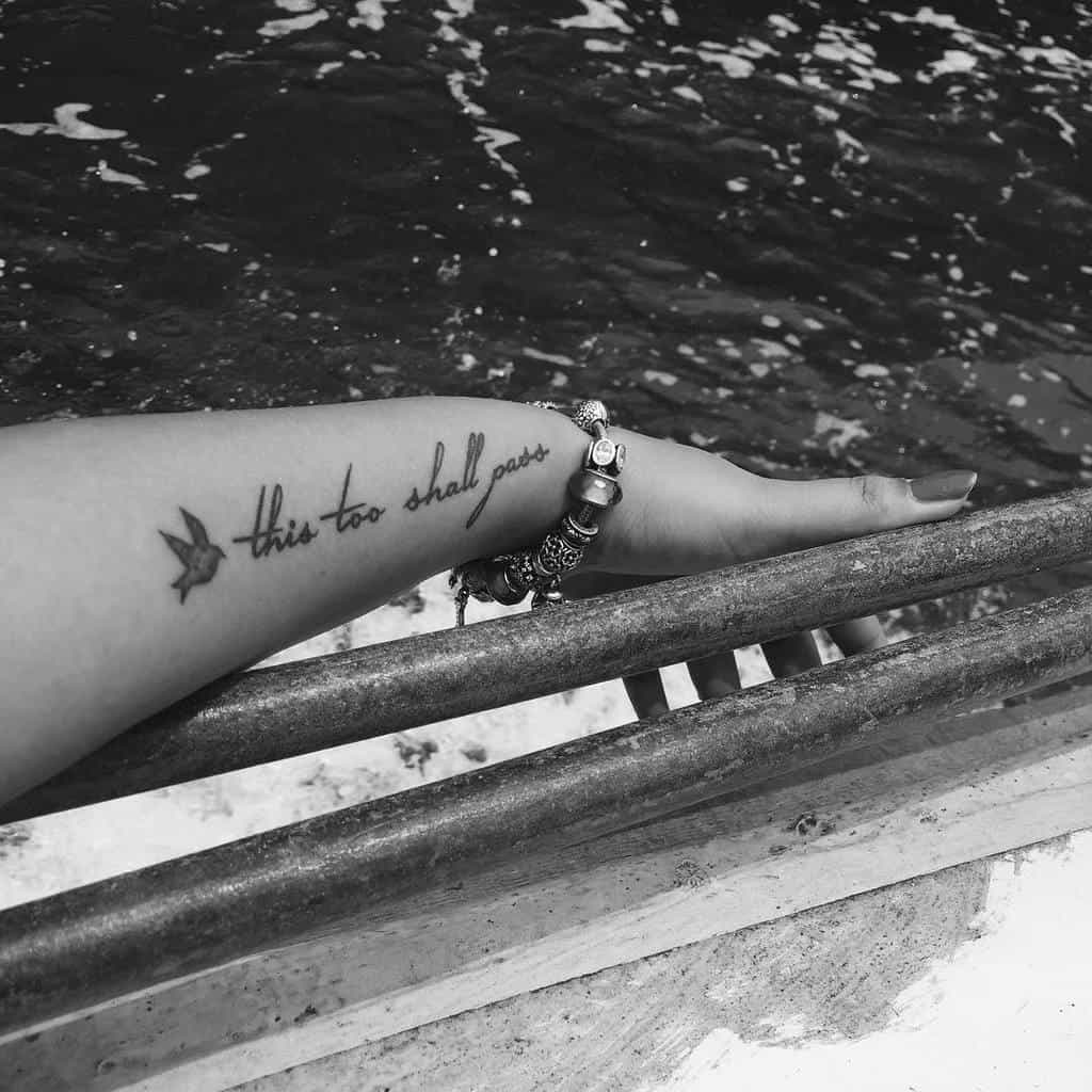 A "This Too Shall Pass" tattoo in cursive with a bird design, on a person's forearm.