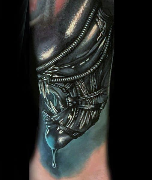 Realistic close-up Xenomorph tattoo featuring sharp teeth, dripping saliva, and detailed tubing in dark metallic tones with a blue glow.