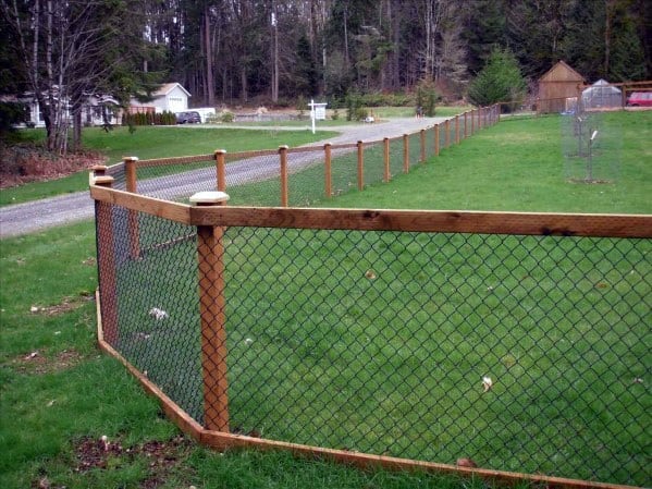 A wooden-post fence encircles a grassy area, with a gravel road and houses visible in the background