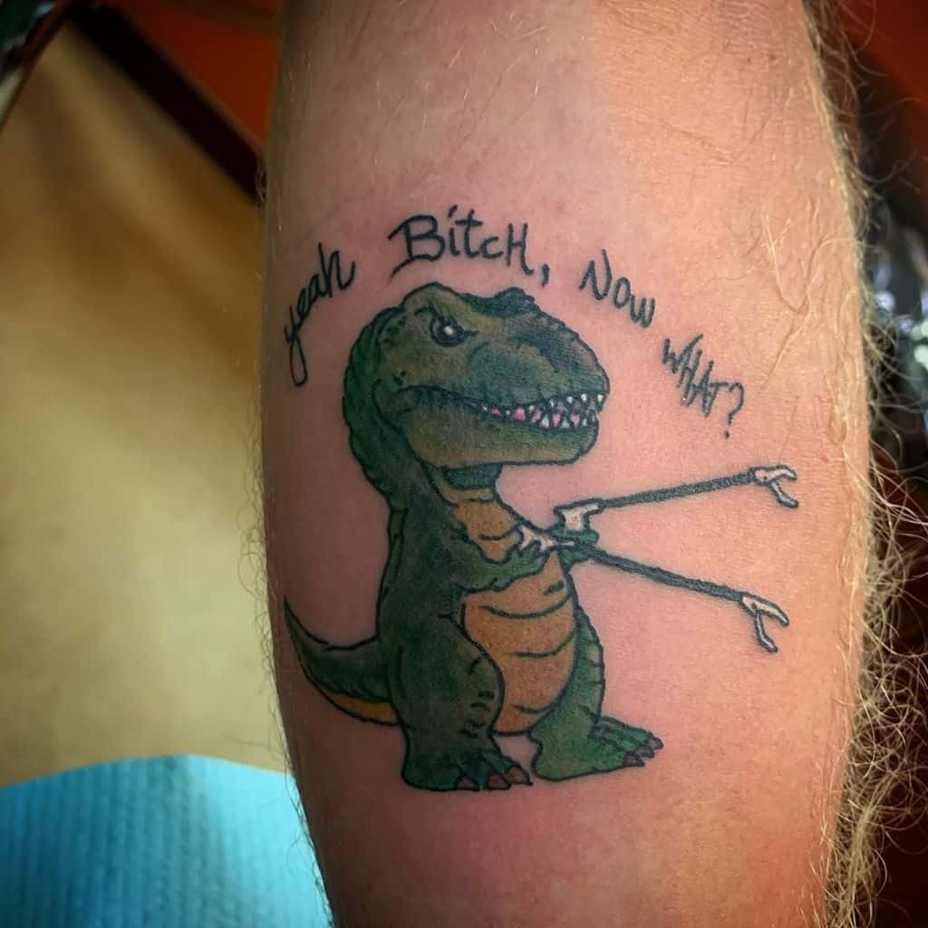 A colorful tattoo of a T-Rex with extended grabber claws and the phrase 'Yeah B****, Now What?' above, blending humor with a cartoonish style.