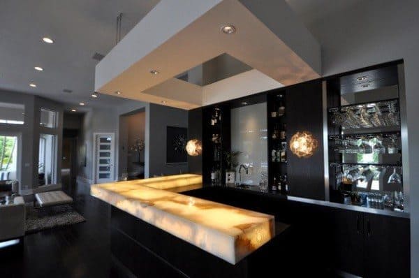 Modern bar with illuminated yellow LED countertop, dark cabinetry, and sleek lighting fixtures.