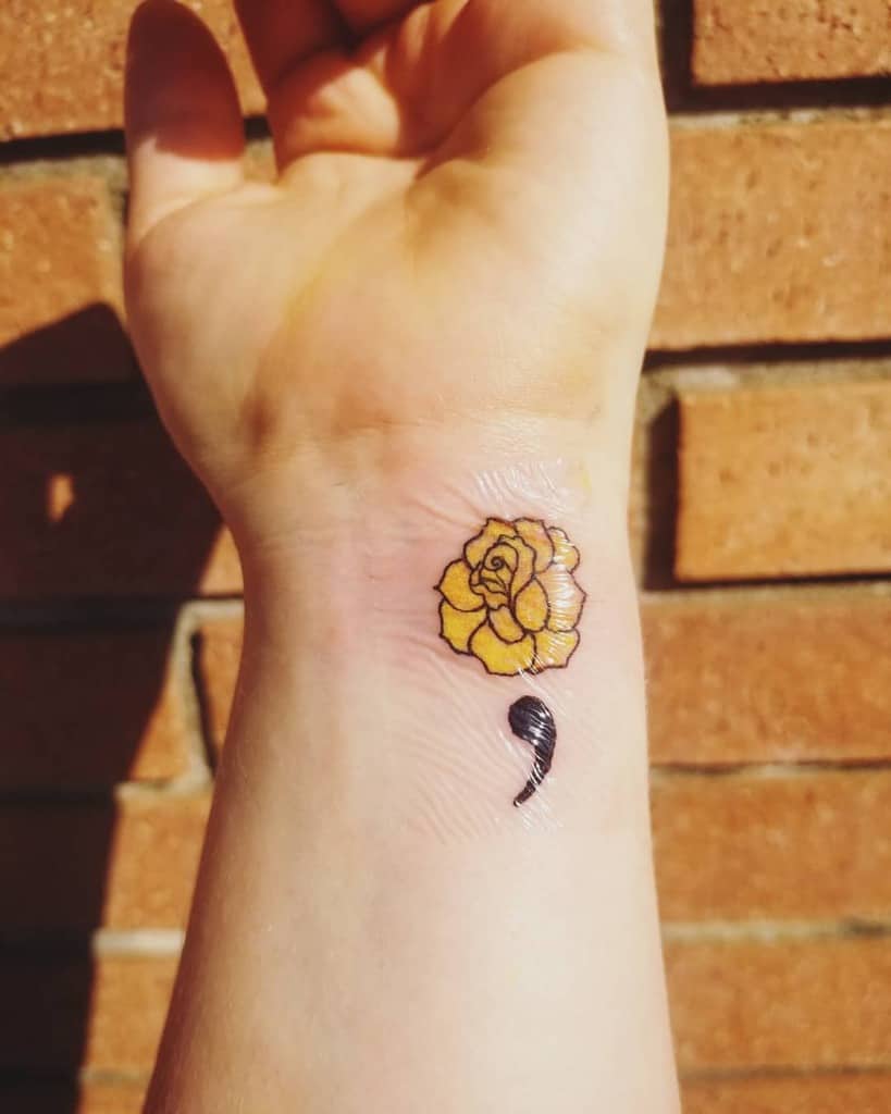 This tattoo features a yellow rose above a semicolon, symbolizing both growth and resilience, often linked to mental health awareness and recovery.