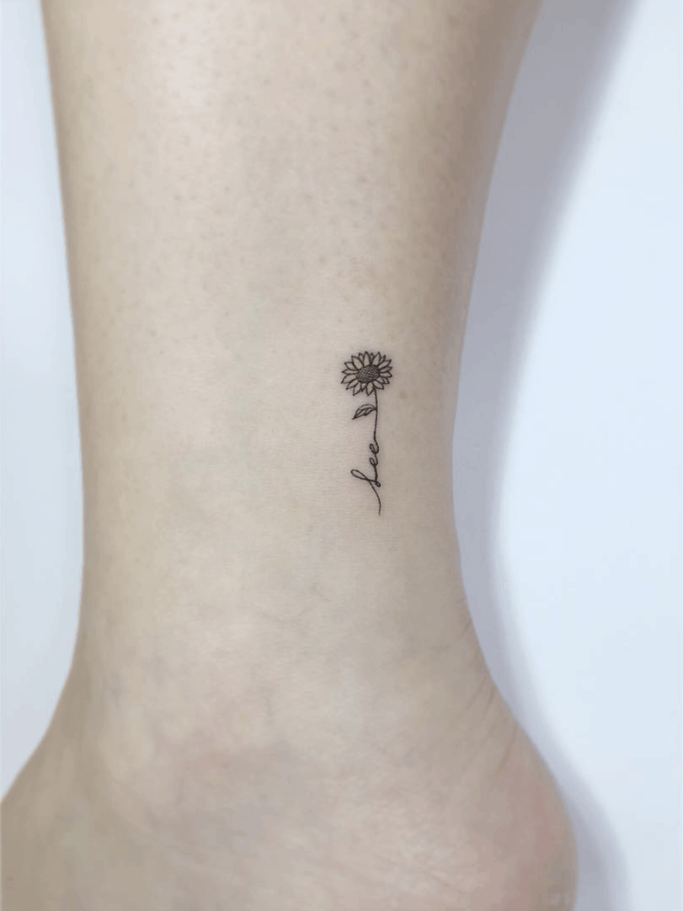 tiny black line tattoo on woman's ankle of a sunflower with the word love in its stem
