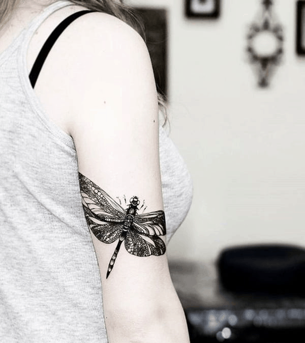 Bold black ink dragonfly tattoo on the upper arm, featuring intricate details and a realistic design with fine shading.