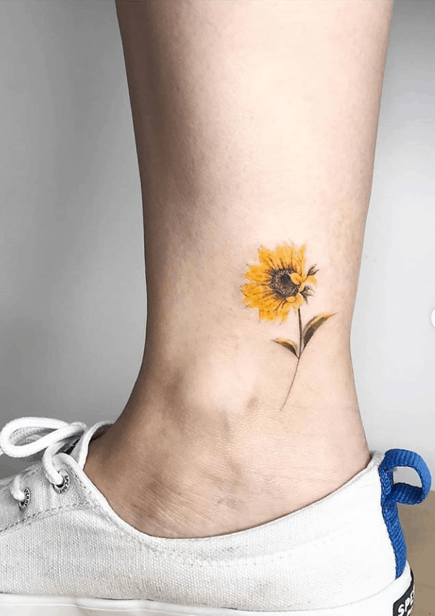 Small, delicate sunflower tattoo on ankle with fine details, soft yellow petals, a brown center, and a thin stem for a minimalist floral design.